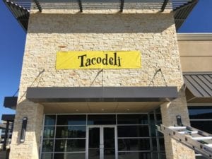 Storefront of Mexican restaurant with large yellow banner that says Tacodeli
