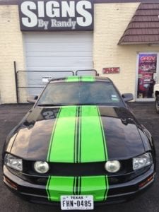 Black Ford Mustang with neon green racing strip vinyl decals in front of Signs by Randy storefront
